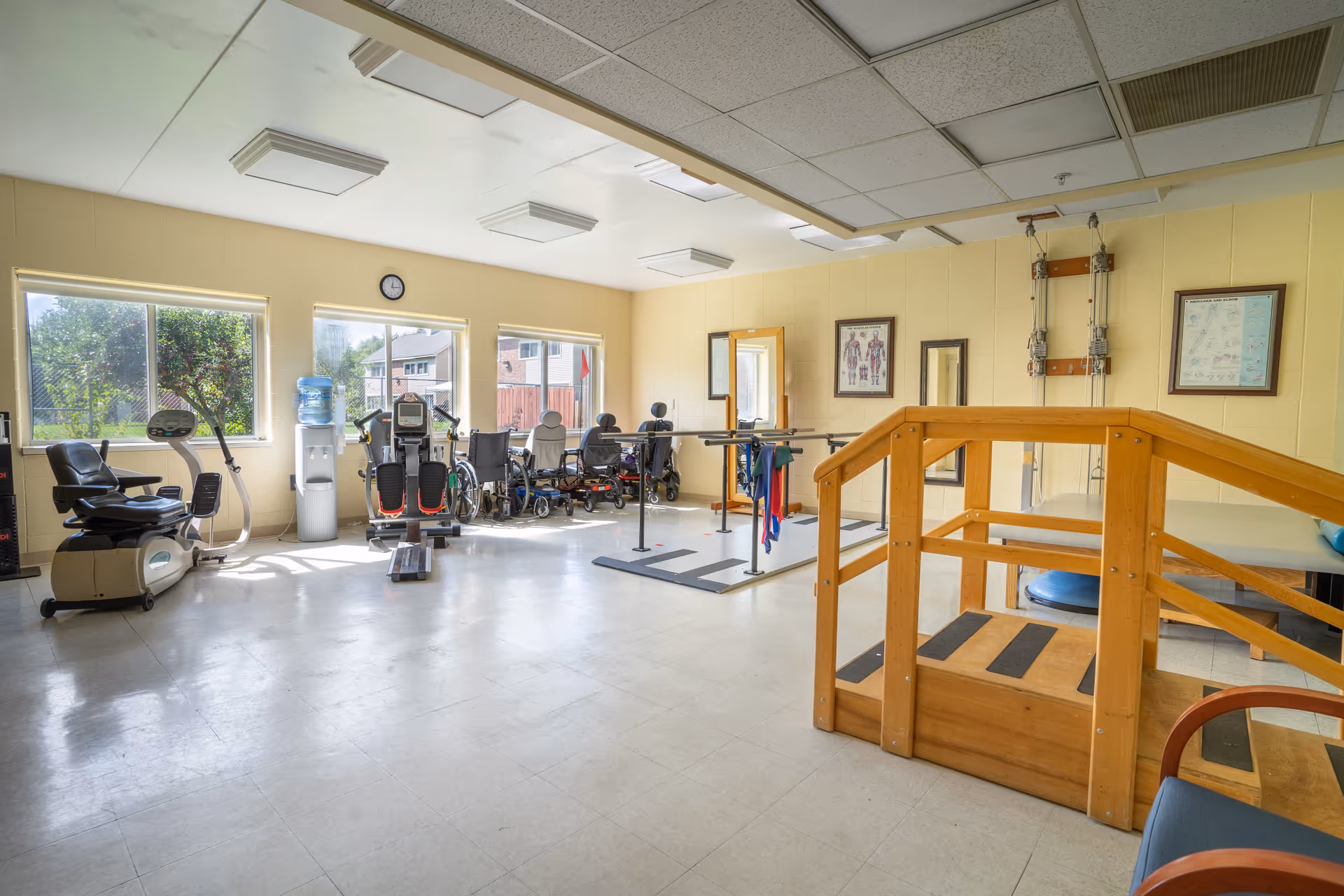 Well-lit interior rehabilitation room with exercise bikes, parallel bars, a wooden practice staircase and other therapy equipment.