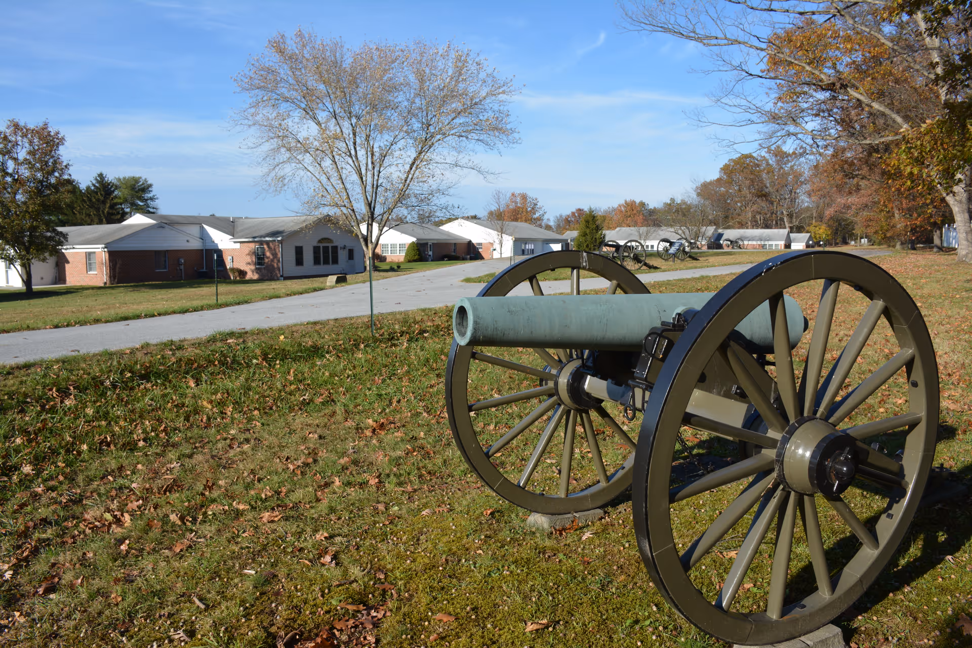 Outdoor scene at SpiriTrust Lutheran, The Village at Gettysburg featuring a historic cannon on a grassy area with a paved road and single-story brick and white buildings in the background under a clear blue sky.