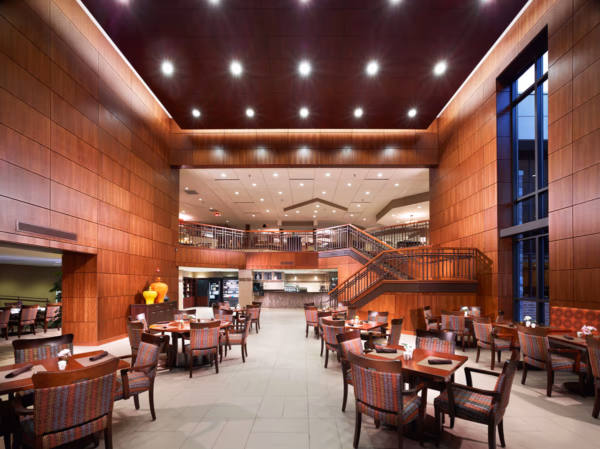 Spacious dining area with multiple wooden tables and chairs featuring patterned upholstery. The room has high ceilings with recessed lighting, wood-paneled walls, and large windows on the right side. A staircase leads to an upper level with additional seating and a kitchen or service area visible in the background.