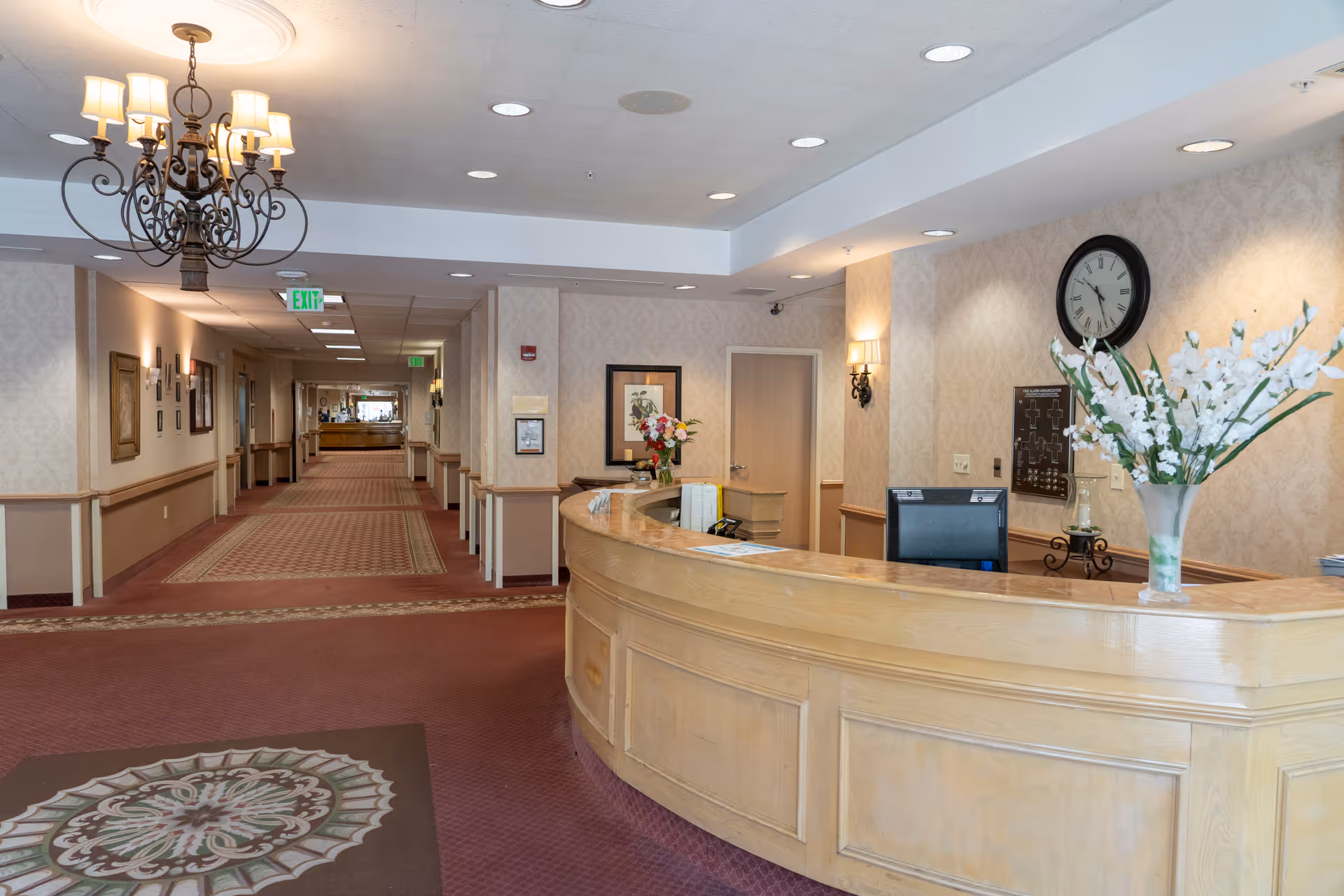 Reception area of Montgomery Village Health Care Center with a curved wooden front desk adorned with a vase of white flowers. Behind the desk is a wall clock and a framed floor plan. The hallway extends into the distance with patterned carpet and framed pictures on the walls. A chandelier hangs from the ceiling near the entrance.