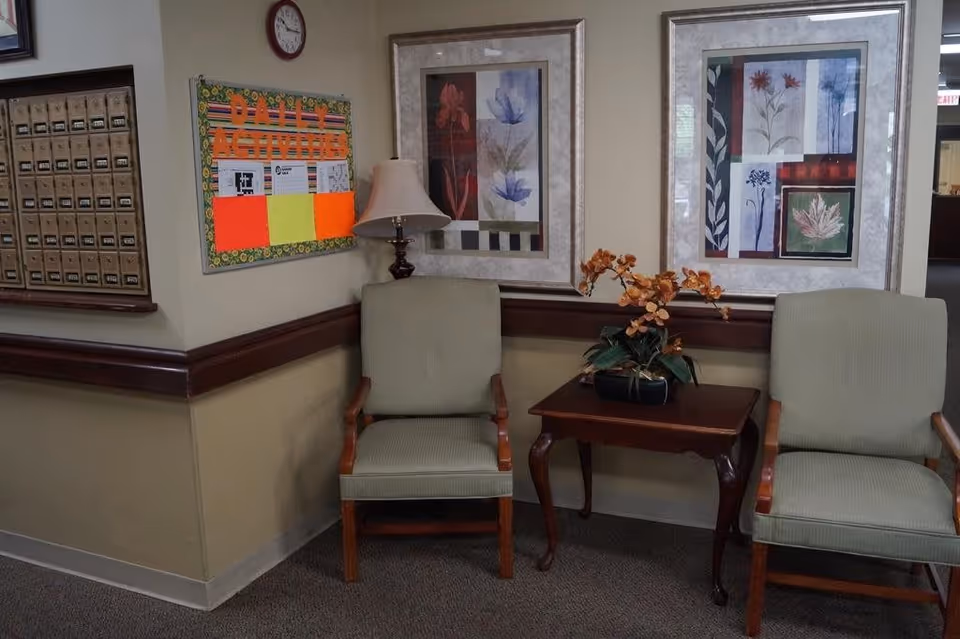 A corner seating area in a senior living facility with two green upholstered chairs separated by a wooden side table holding a potted plant with orange flowers. Behind the chairs are two framed floral artworks on the wall. To the left, there is a bulletin board with colorful papers and a clock above it, as well as a set of mailboxes mounted on the wall.