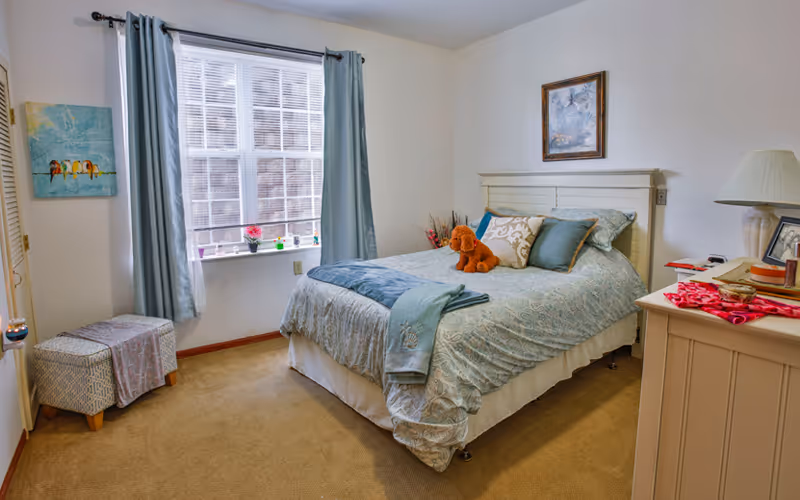 A cozy bedroom with a neatly made bed featuring blue and white bedding, decorative pillows, and a stuffed dog toy on top. There is a window with light blue curtains letting in natural light, a small ottoman covered with a patterned cloth near the window, a dresser with a lamp and framed photos, and a painting hanging above the bed.