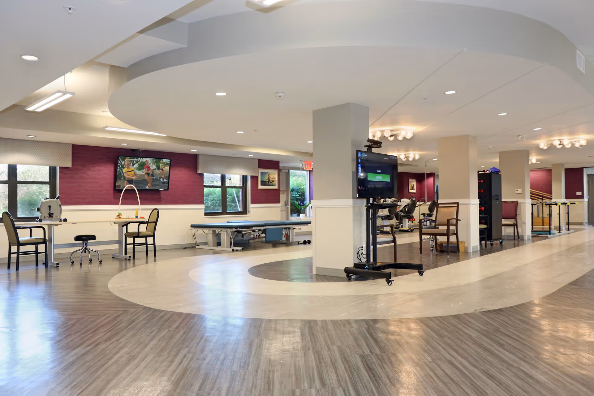 Spacious rehabilitation and therapy room with exercise equipment, therapy tables, chairs, and a TV mounted on a mobile stand. The room has large windows with natural light, purple accent walls, and a circular ceiling design with recessed lighting.