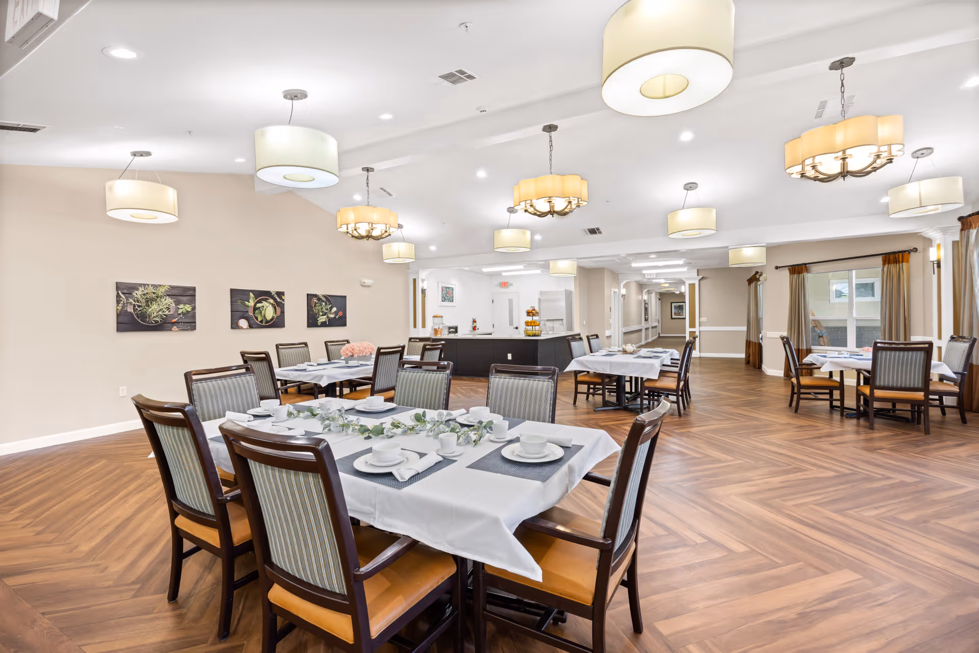 A spacious dining room in a senior living facility with several tables covered with white tablecloths, set with plates, cups, and silverware. The room features wooden flooring, multiple modern ceiling lights, and framed botanical artwork on the walls. Large windows with curtains allow natural light to enter the room.