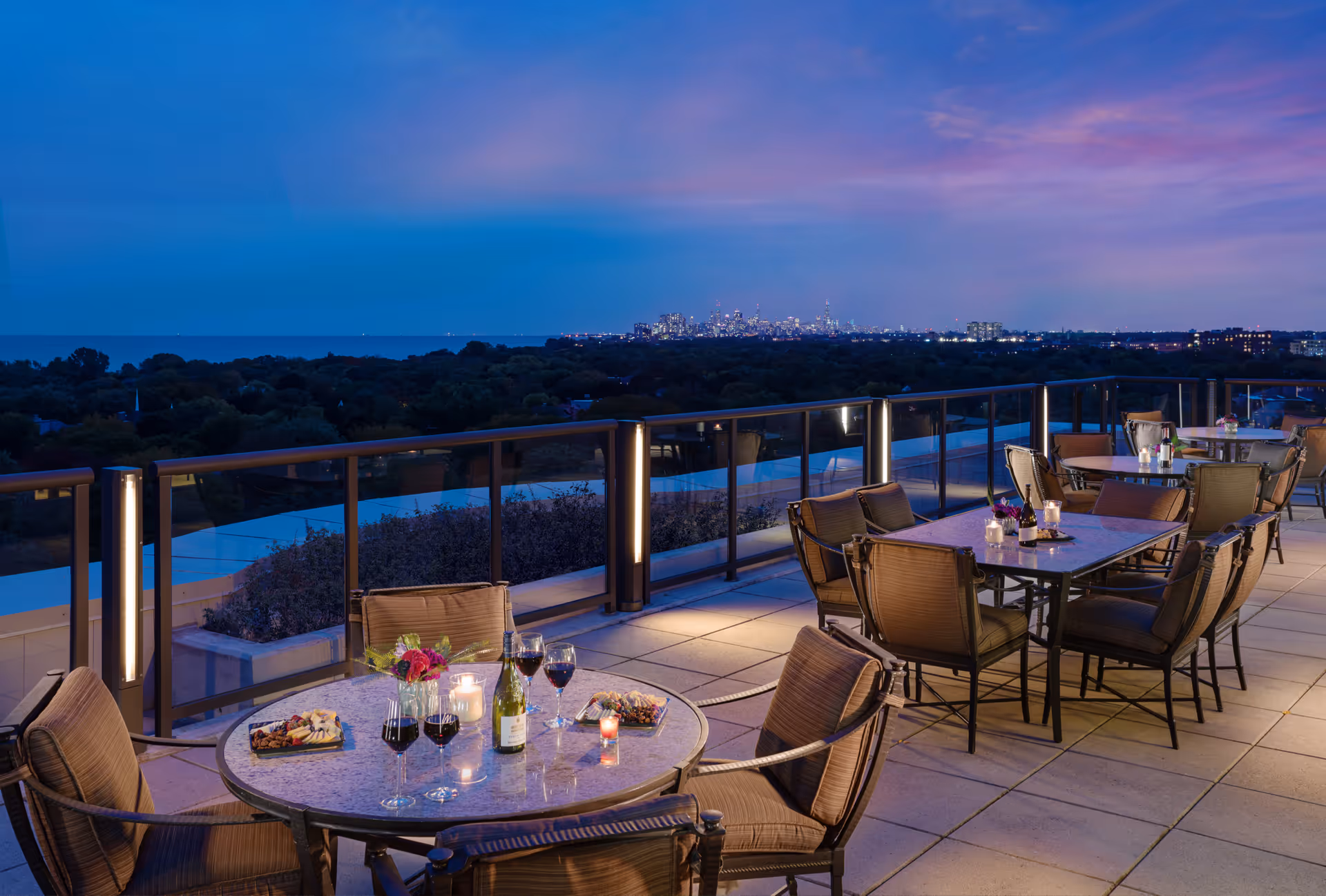 Outdoor patio area at dusk with tables and chairs set for dining, featuring wine glasses, a bottle of wine, candles, and flowers. The patio overlooks a scenic view of trees and a distant city skyline under a purple and blue sky.