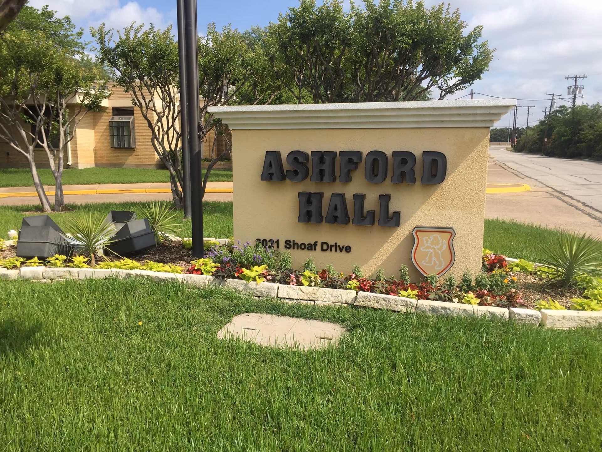 Outdoor view of a sign for Ashford Hall Nursing and Rehabilitation located at 2031 Shoaf Drive, surrounded by green grass, small plants, flowers, and trees with a building and street in the background under a partly cloudy sky.