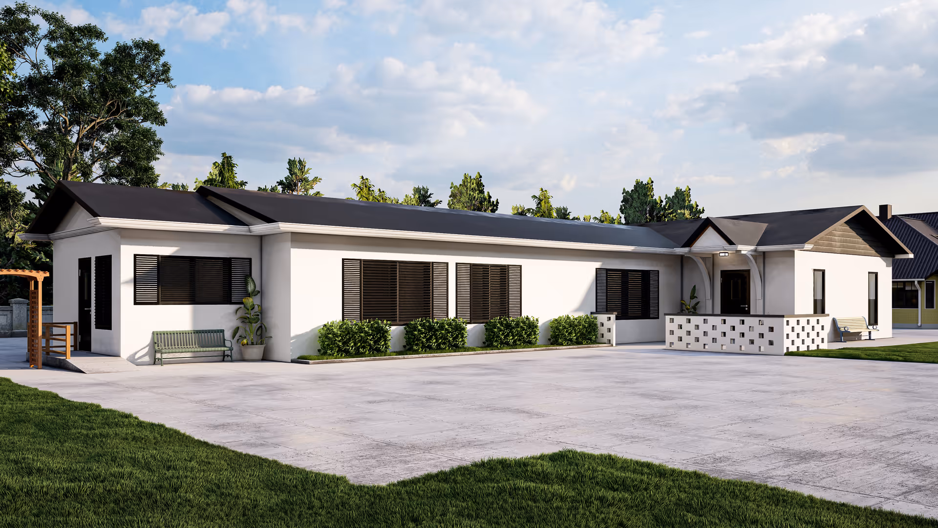 Exterior view of a single-story white building with black window shutters and a dark roof, surrounded by green grass and trees under a partly cloudy sky. The building has a paved area in front and benches near the entrances.