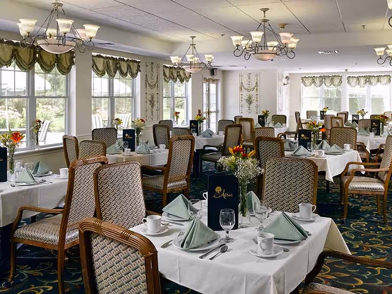 A dining room with multiple tables set for a meal, each table covered with a white tablecloth, green folded napkins, cups, glasses, and silverware. The chairs have patterned upholstery and wooden frames. The room has large windows with green valances, chandeliers hanging from the ceiling, and floral centerpieces on each table.
