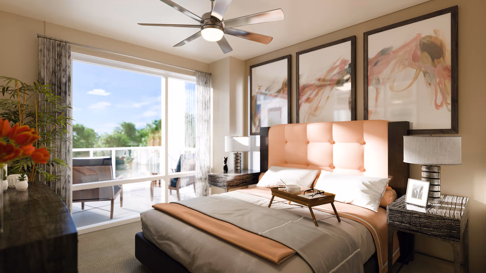 A bright bedroom with a large window and glass door leading to a balcony with outdoor seating. The room features a bed with a peach-colored tufted headboard, white and peach bedding, a wooden tray with a bowl and glasses, two bedside tables with lamps, abstract artwork above the bed, a ceiling fan, and a dresser with plants and flowers.