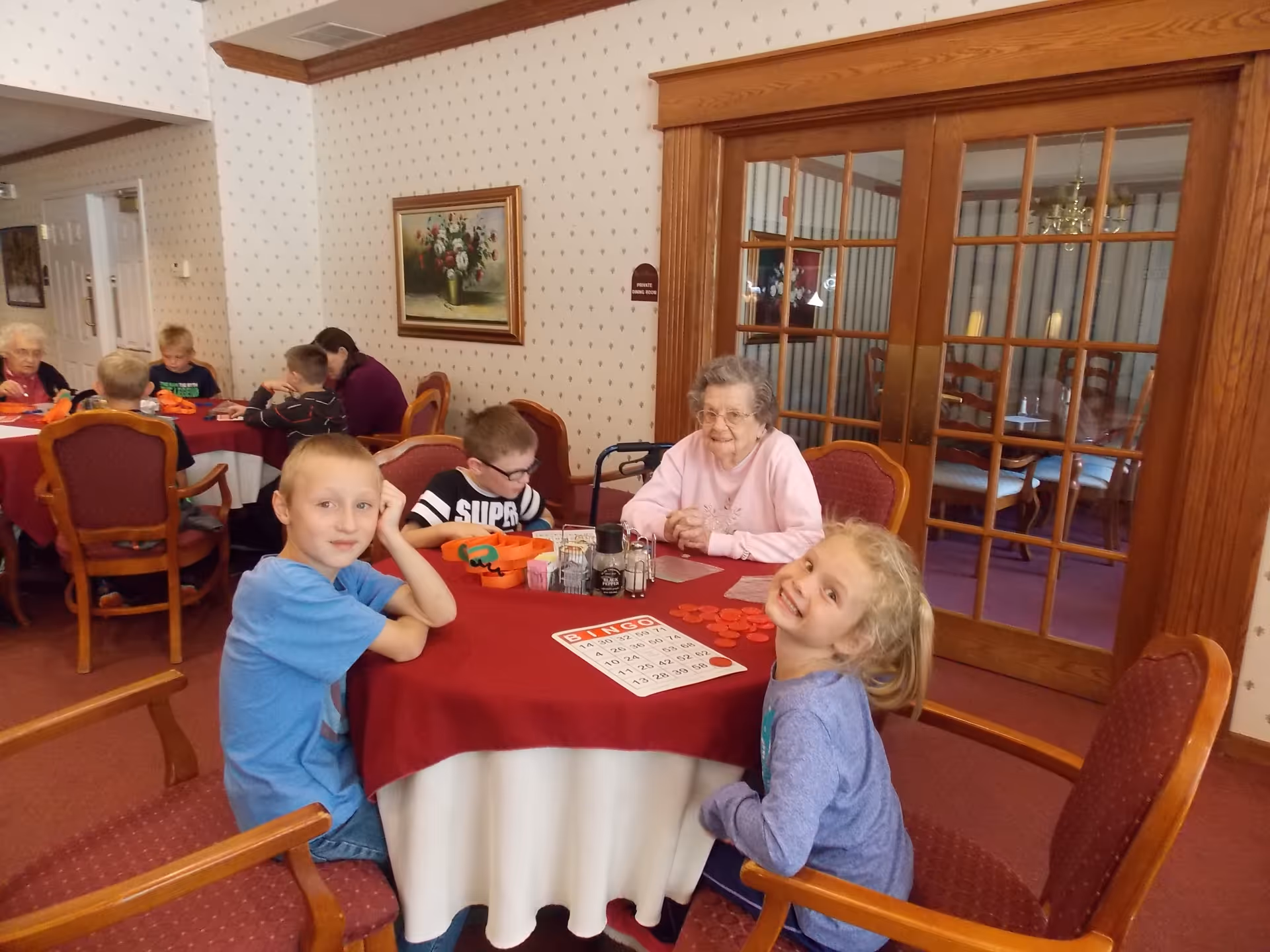 An elderly woman and several children sitting around tables covered with red and white tablecloths in a room with patterned wallpaper and wooden framed glass doors. They appear to be playing bingo, with bingo cards and red chips on the tables. A painting of flowers hangs on the wall.
