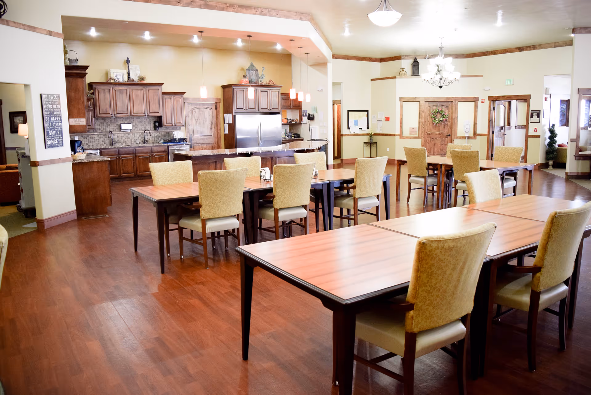 A spacious dining area in an assisted living facility featuring multiple wooden tables with cushioned chairs. The room has hardwood floors, warm lighting, and a kitchen area with wooden cabinets, a refrigerator, and a countertop in the background. The walls are light-colored with wooden trim, and there is a decorative wreath on a door at the far end.