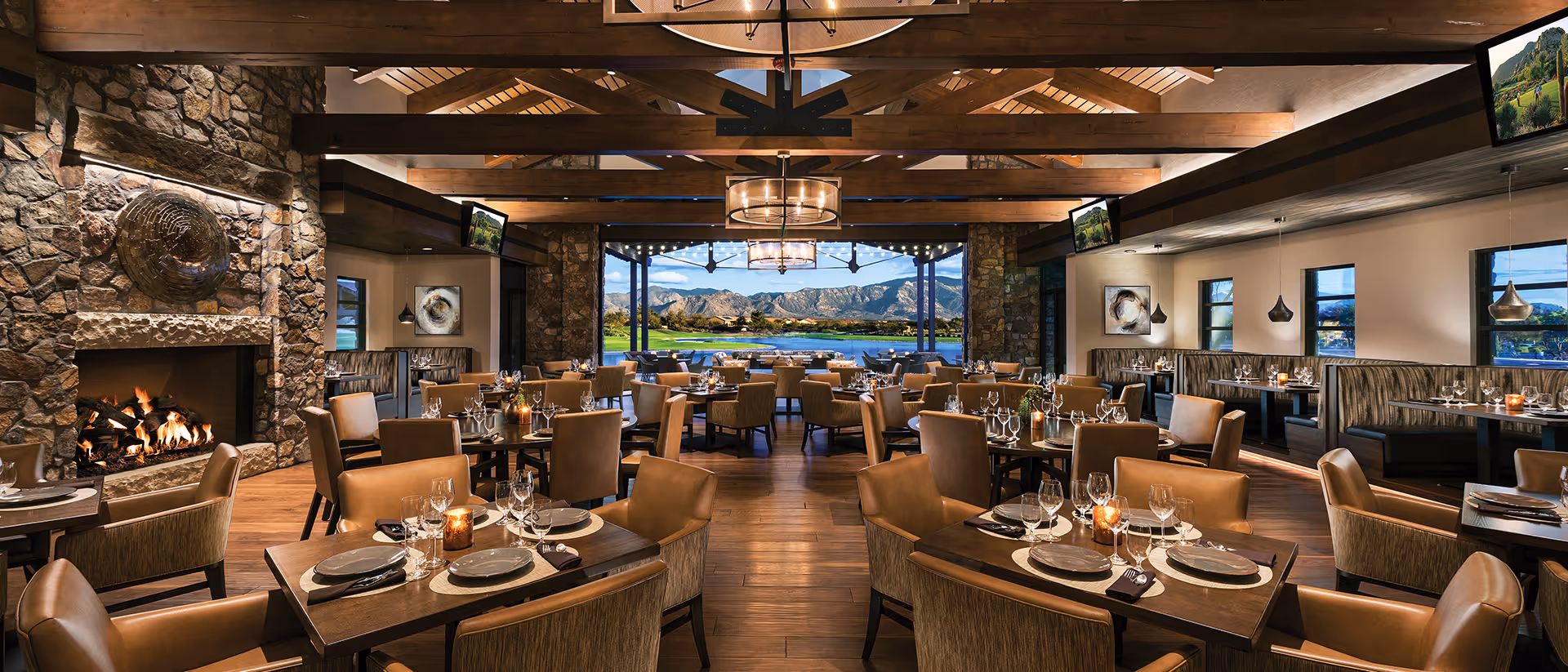 A spacious, elegant dining room with wooden beams on the ceiling, stone walls, and a large stone fireplace on the left. The room is filled with tables set with plates, glasses, and candles. Large windows and an open wall at the far end offer a scenic view of mountains and a lake outside. The lighting is warm and inviting, with pendant lights and chandeliers.
