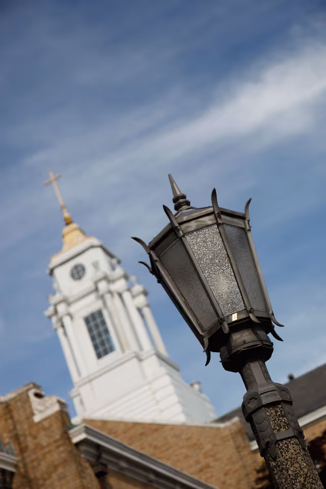 Close-up view of a decorative black street lamp with a textured glass design, set against the backdrop of a white church steeple topped with a golden cross and a blue sky with wispy clouds.