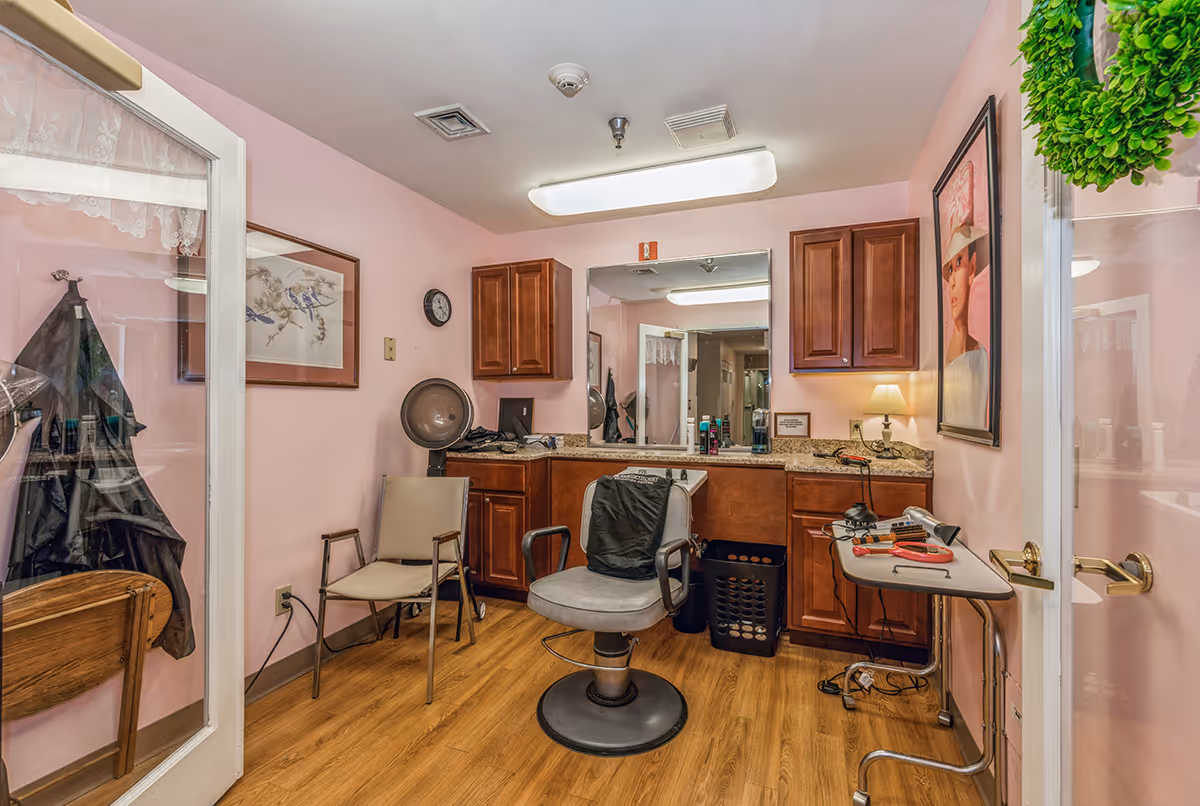 Interior view of a hair salon room with pink walls and wooden flooring. The room features a salon chair in the center, a hair dryer hood, a small table with hair styling tools, wooden cabinets, a large mirror, and framed artwork on the walls. A green wreath hangs on the glass door to the right.