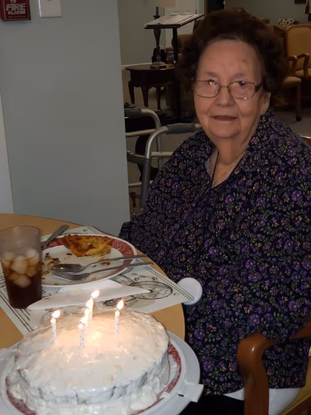 An elderly woman with glasses and curly hair sits at a table in a dining area. She is wearing a dark floral blouse and smiling slightly. On the table in front of her is a white frosted birthday cake with four lit candles, a plate with a partially eaten slice of pizza, a fork and knife, and a glass of iced tea. In the background, there is a walker and some furniture.