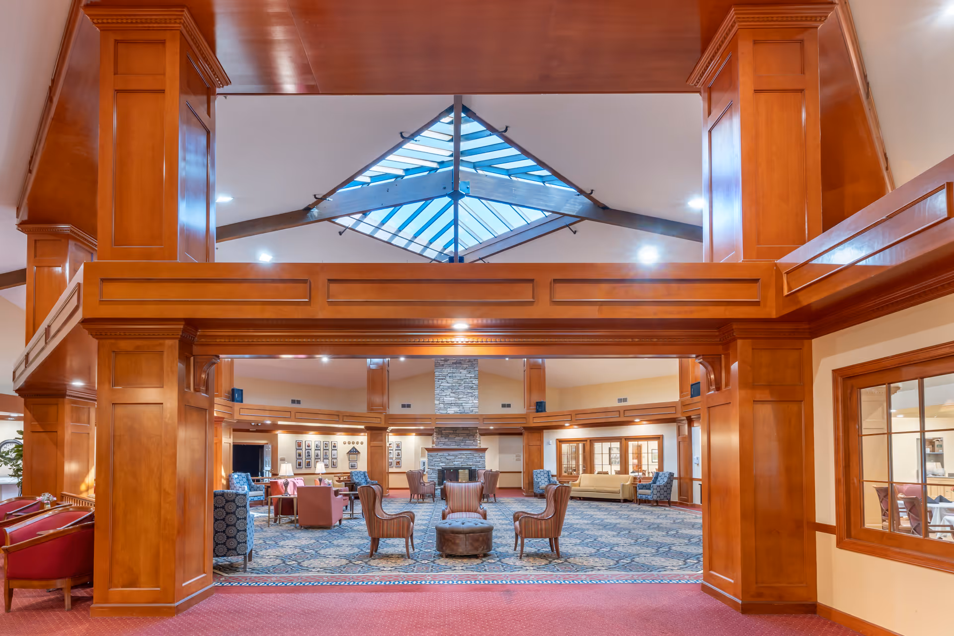 Spacious senior living facility common area with high wooden beams and a large skylight. The room features a patterned carpet, multiple seating arrangements including armchairs and sofas, and a stone fireplace at the far end. Warm lighting and wood paneling create a cozy atmosphere.