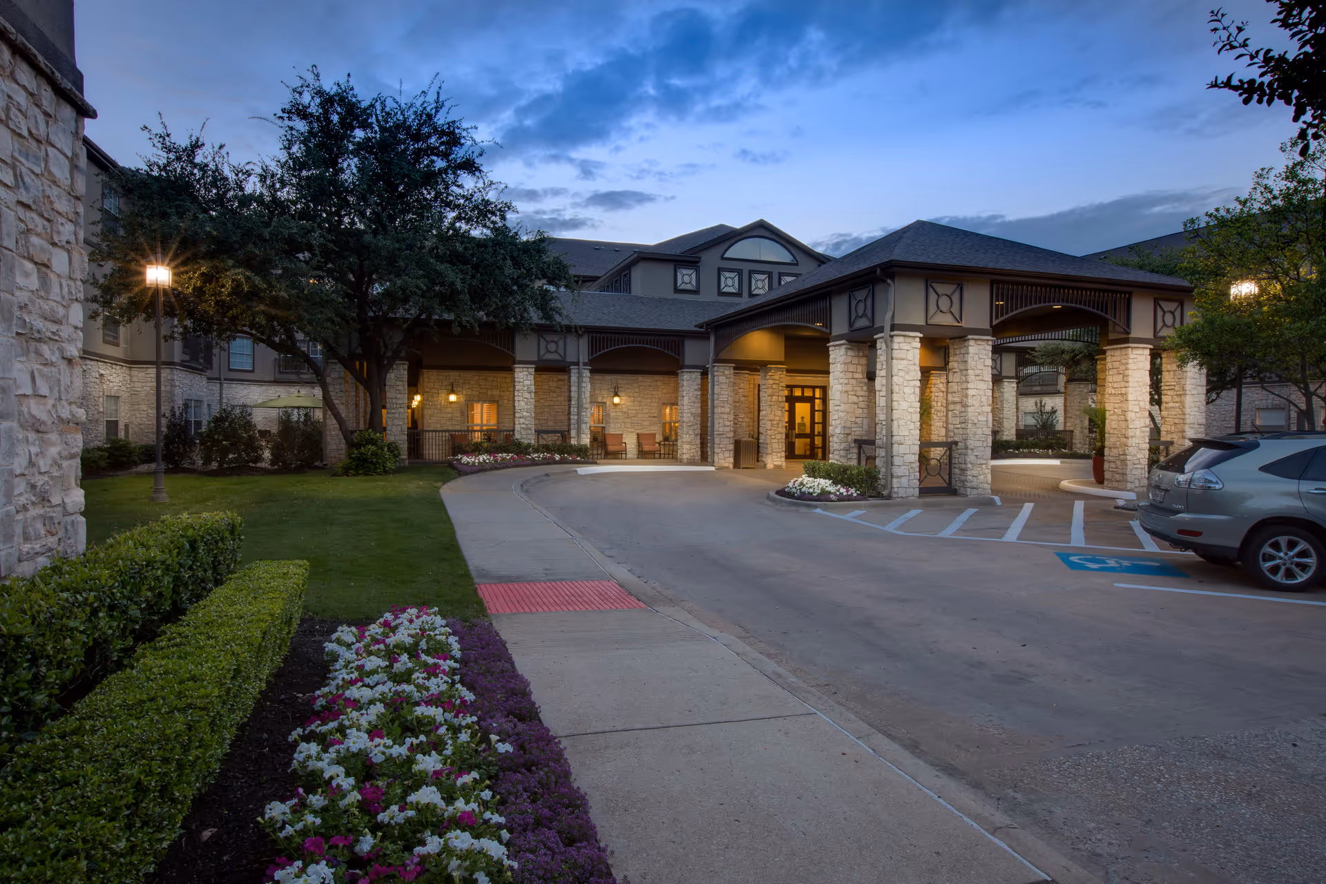 Entrance of The Reserve at North Dallas Assisted & Senior Living facility during dusk, showing a covered driveway with stone pillars, landscaped flower beds, green bushes, a tree, and parked cars near a handicapped parking space.