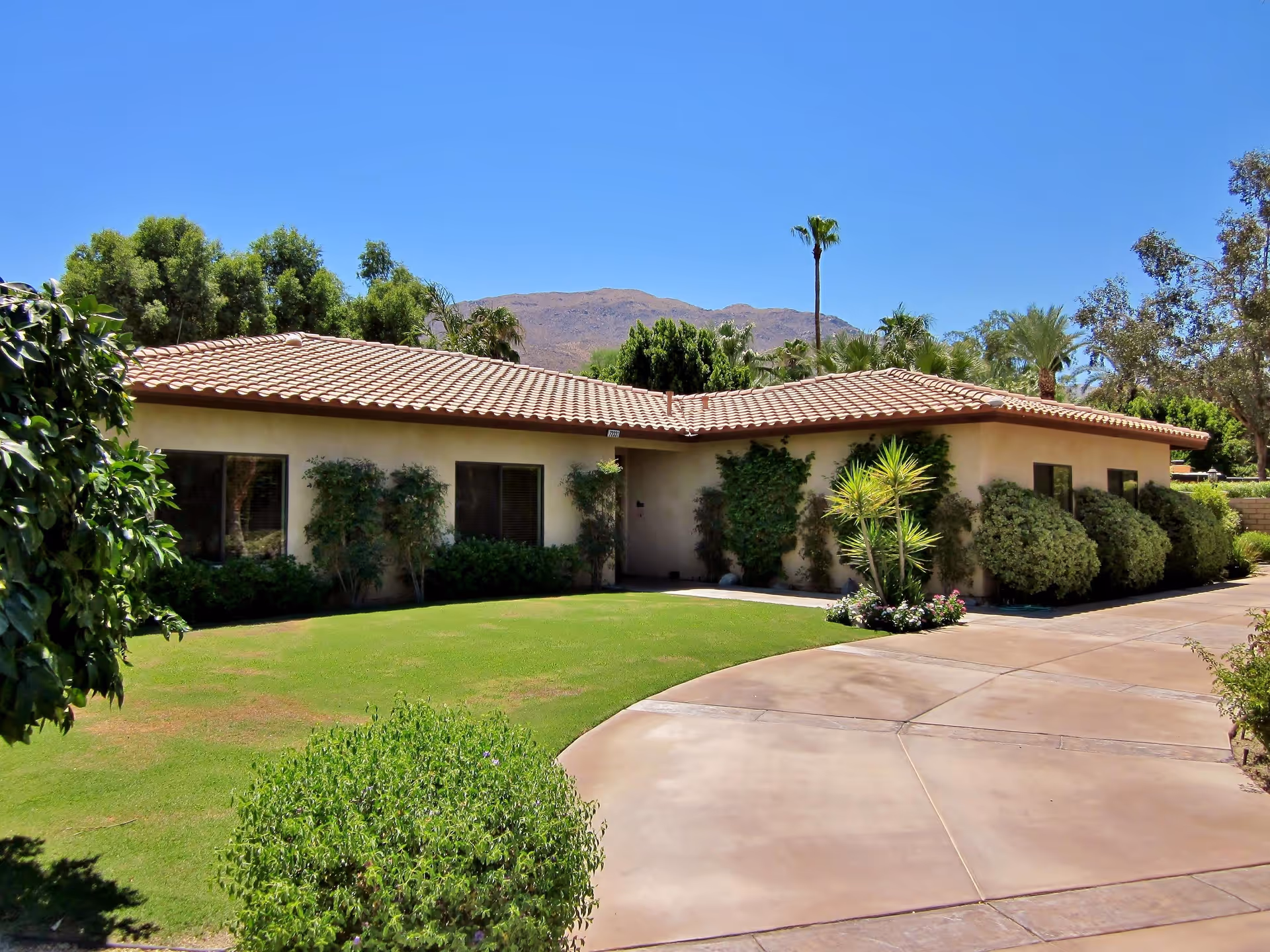 Single-story building with a tiled roof surrounded by green bushes and trees, a well-maintained lawn, and a concrete driveway under a clear blue sky with mountains in the background.