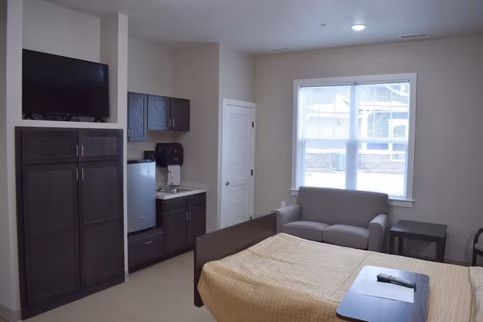 A senior living facility room with a bed covered in a beige quilt, a small sofa near a window with blinds, a side table, and a kitchenette area with dark wood cabinets, a mini refrigerator, and a small sink. A flat-screen TV is mounted above a cabinet on the left wall.