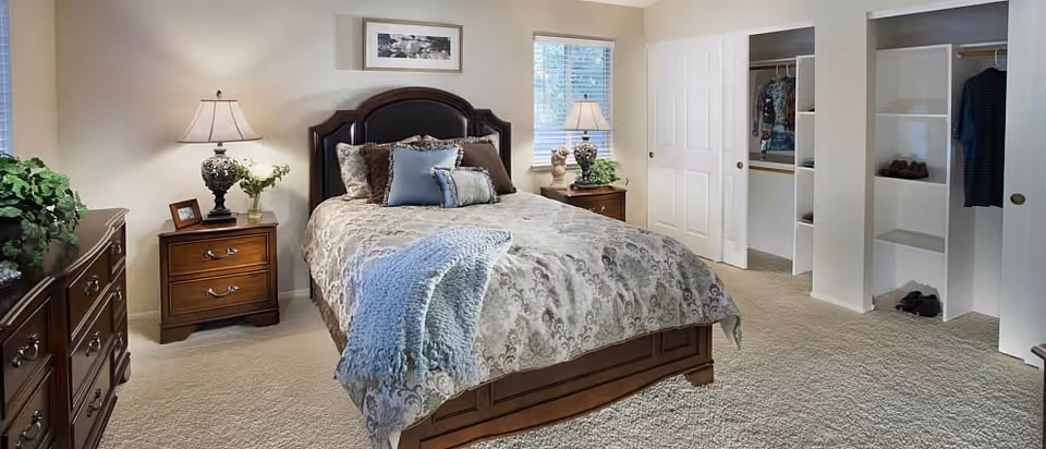 Well-lit bedroom with a bed, wooden nightstands and dresser, bedside lamps, and open closets along one wall.