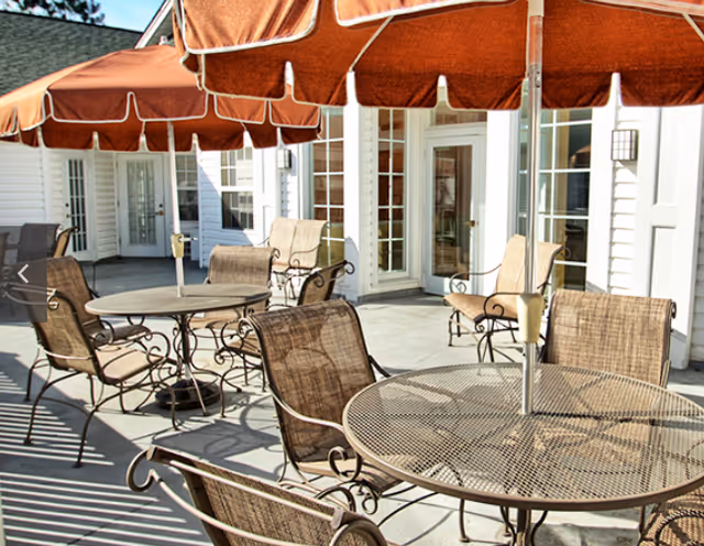 Outdoor patio area with round metal tables and brown cushioned chairs under large rust-colored umbrellas, adjacent to a white building with multiple glass doors and windows.