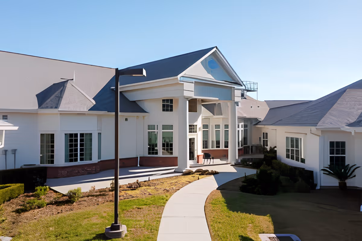 Exterior view of SummerHouse Ashton Manor, showing a large white building with multiple windows, a peaked roof, and a covered entrance supported by columns. A curved sidewalk leads to the entrance, surrounded by grass and landscaped areas under a clear blue sky.