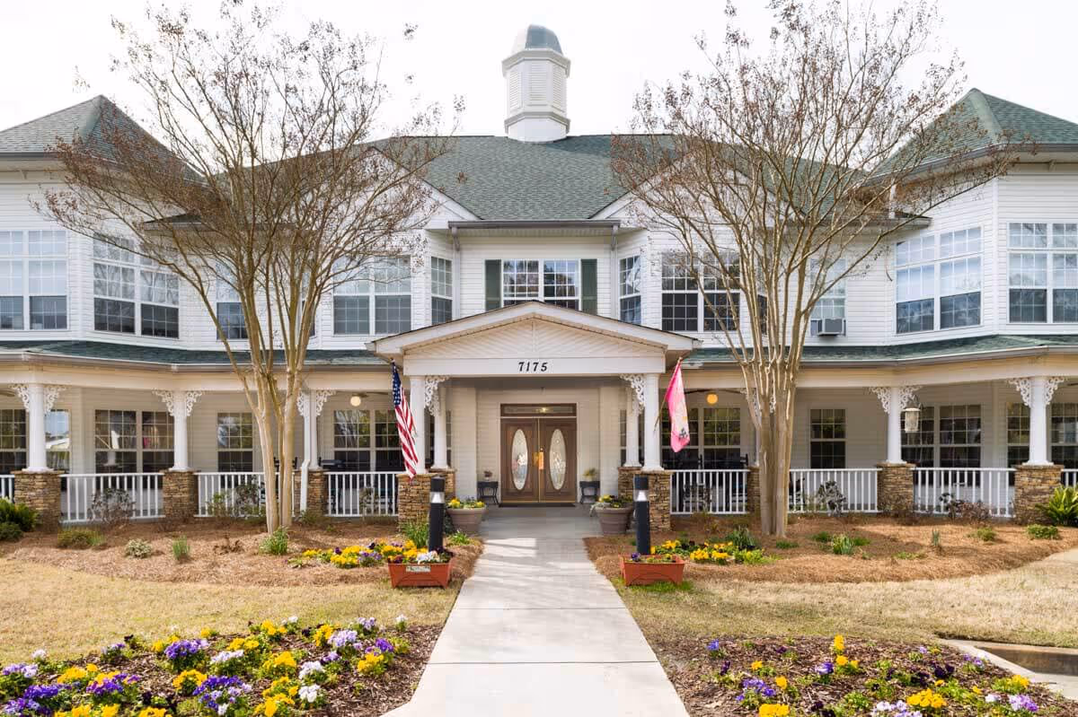 Front exterior view of a large two-story white building with green roof shingles, a covered entrance with double doors, two flags on either side of the entrance, and landscaped flower beds with colorful flowers in the foreground.