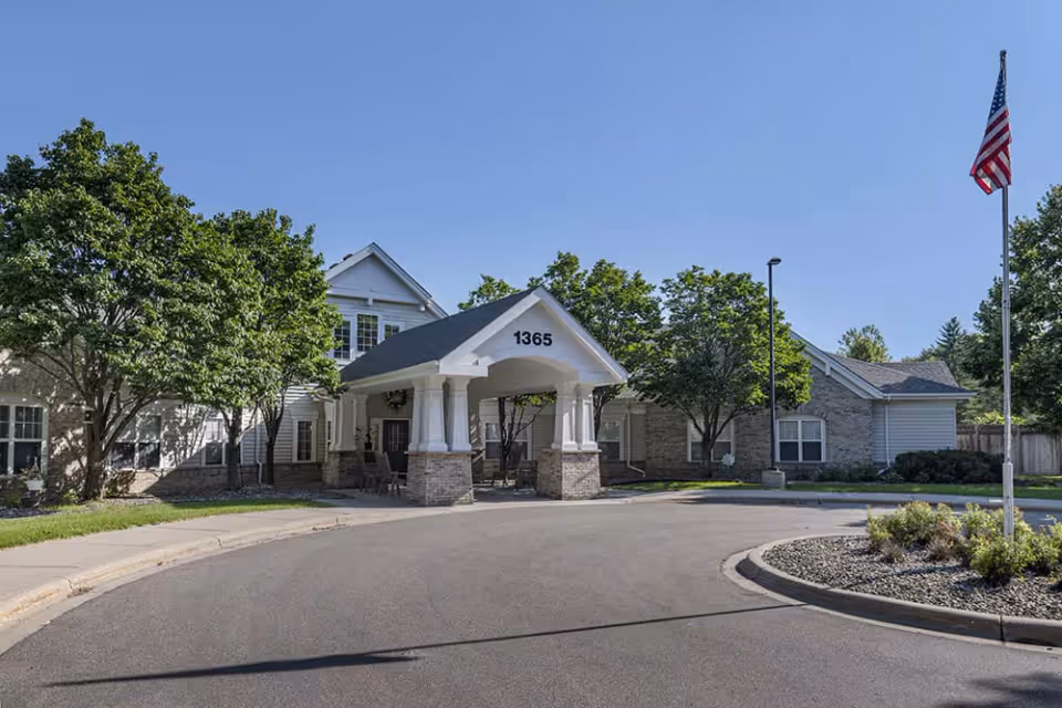 Exterior view of a senior living facility entrance with a covered drop-off area supported by white columns and the number 1365 displayed above. The building is surrounded by trees and landscaping, with an American flag on a flagpole to the right.