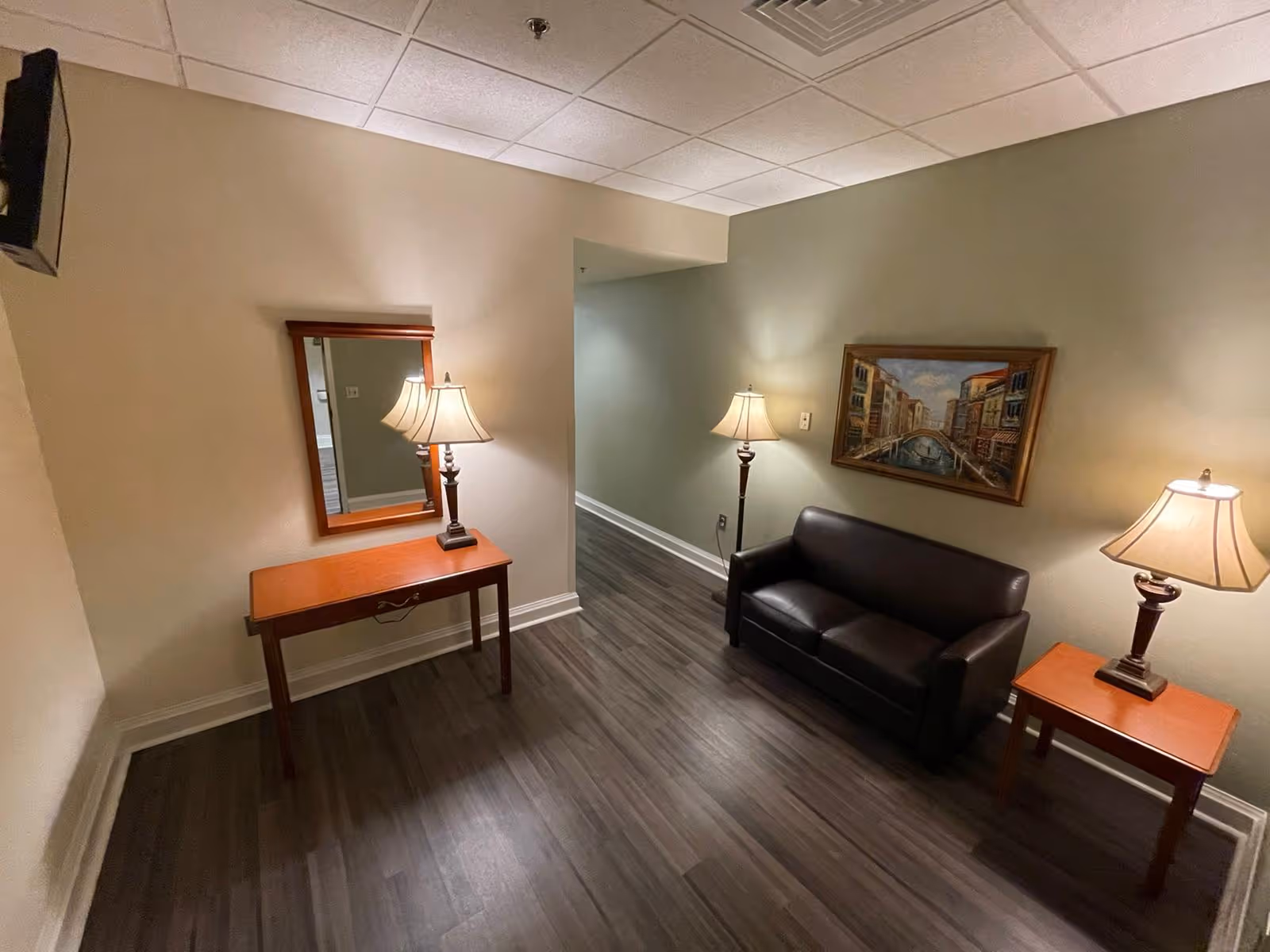 A small interior room with dark wood flooring, beige walls, and a white ceiling. The room contains a black leather loveseat, two wooden side tables each with a lamp, a wooden console table with a lamp and a mirror above it, and a framed painting of a canal scene on the wall.