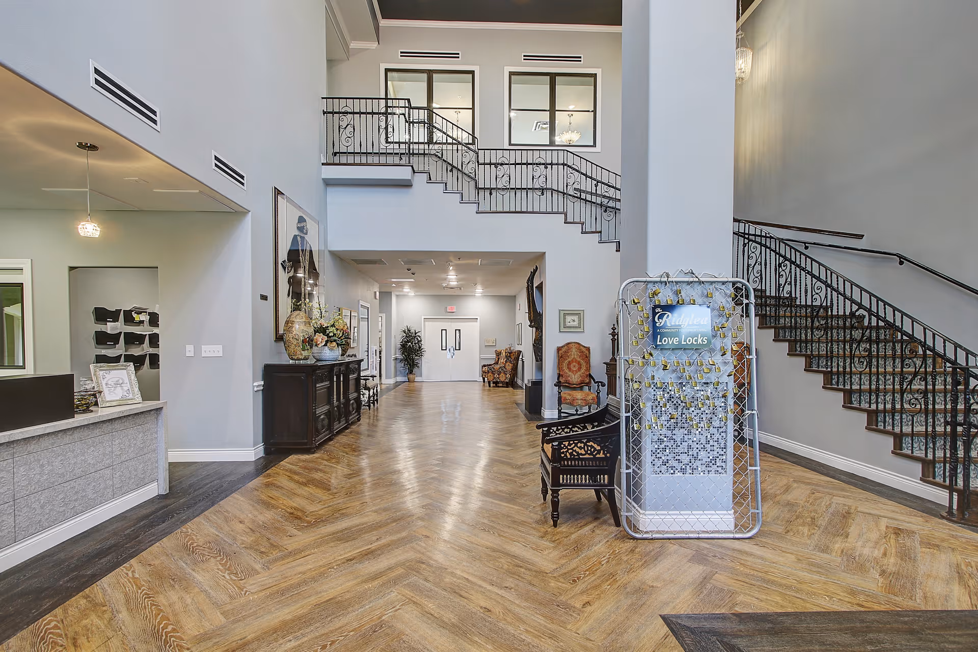 Spacious senior living facility lobby with a high ceiling, decorative staircase with wrought iron railing, wooden herringbone floor, reception desk on the left, and a display stand labeled 'Ridglea Love Locks' in the center. The area is decorated with chairs, plants, and artwork, creating a welcoming atmosphere.
