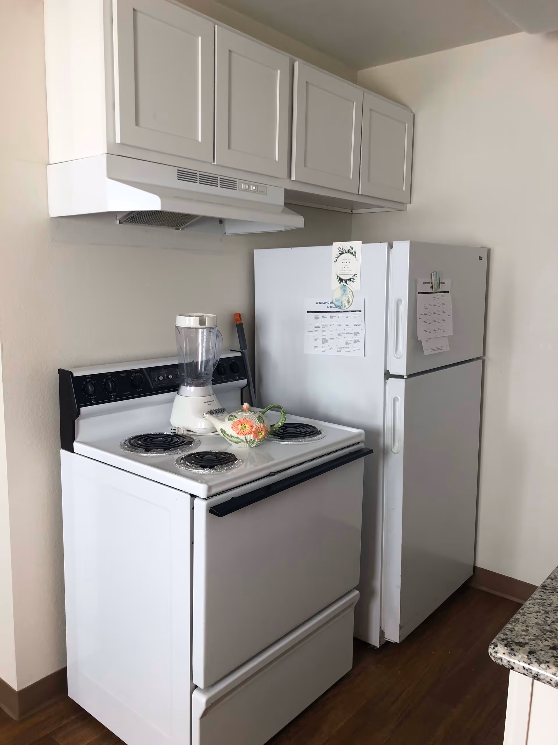A small kitchen area with a white electric stove, a white blender, and a decorative teapot on the stove. Above the stove are white cabinets and a white range hood. Next to the stove is a white refrigerator with papers and notes attached to the door. The floor is wooden and part of a granite countertop is visible on the right.