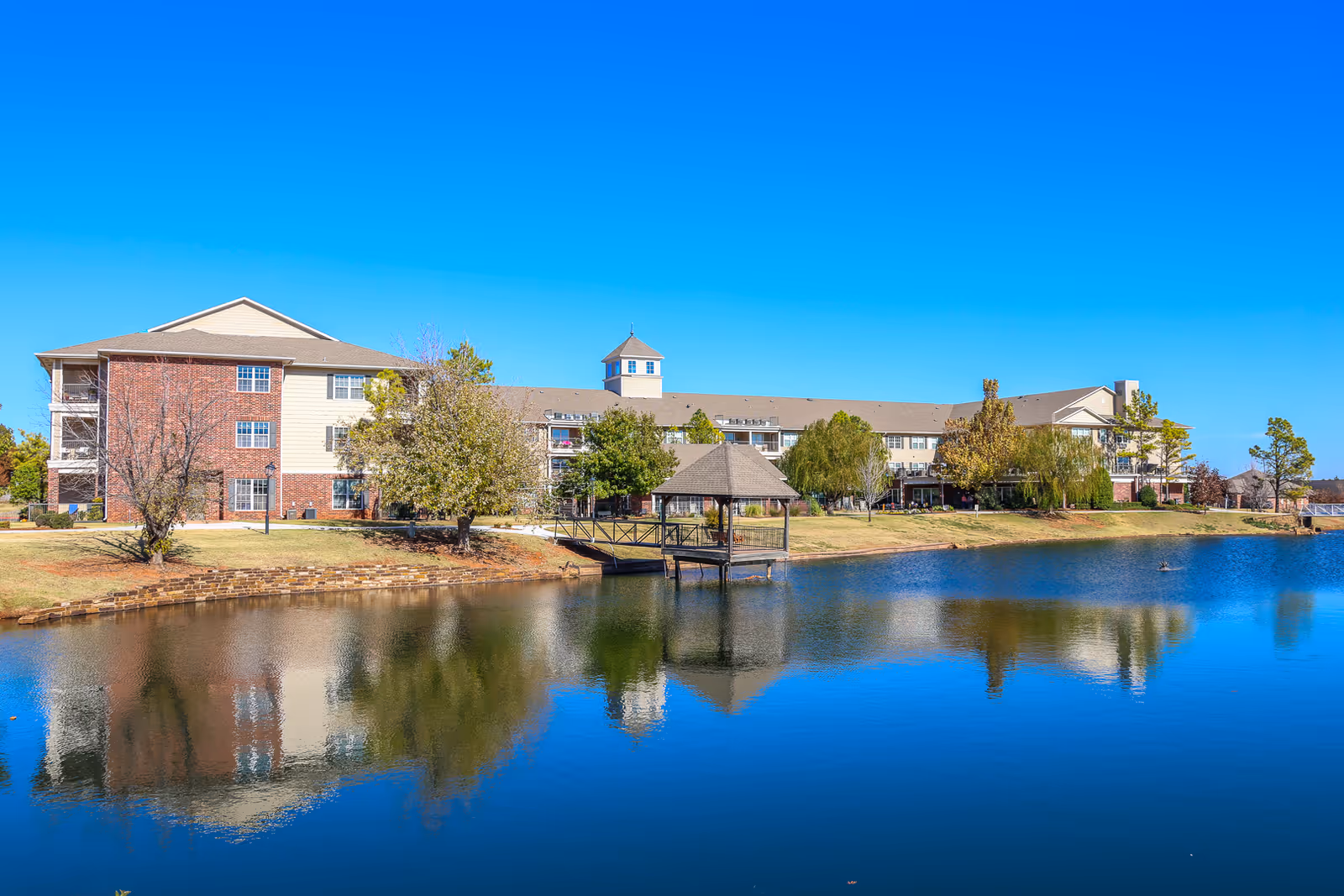 A large senior living facility building with a mix of brick and beige siding, situated beside a calm pond with clear blue water reflecting the building and surrounding trees. There is a small wooden gazebo on the pond connected by a walkway. The sky is clear and bright blue.
