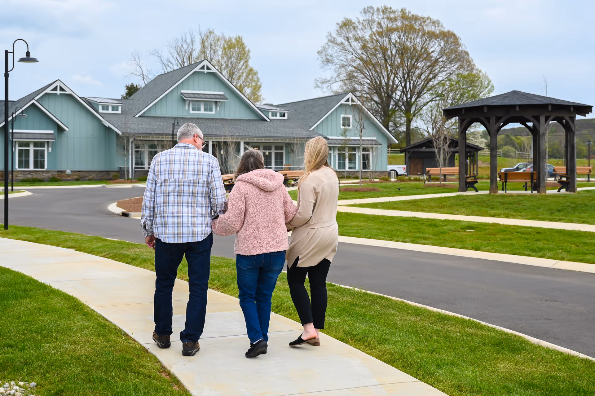 Three people walking on a sidewalk in a senior living community. Two women and one man are seen from behind, walking arm in arm towards a large light blue building with a gray roof. The area is landscaped with green grass, trees, benches, and a wooden gazebo.