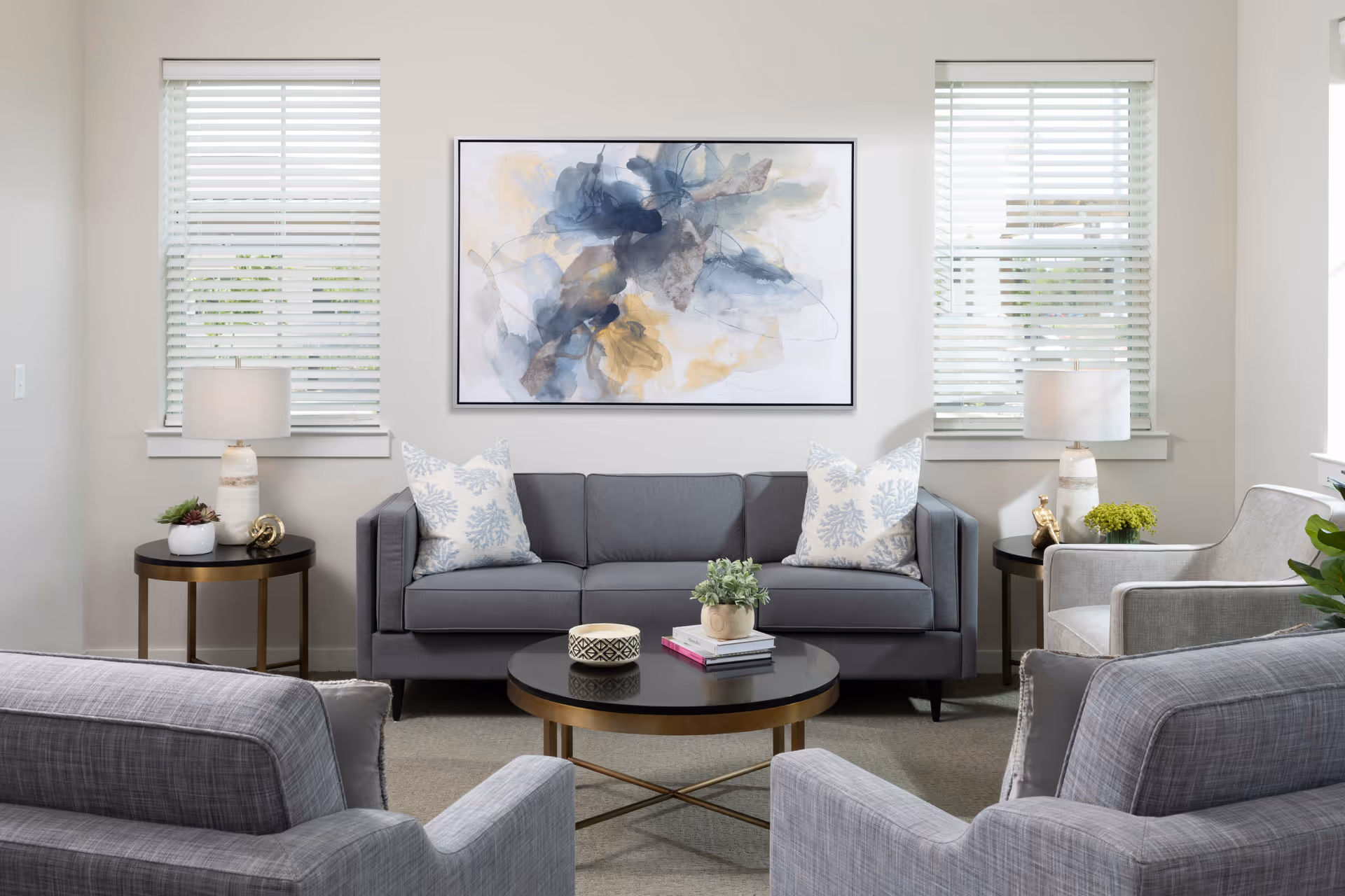 A modern living room with a gray sofa adorned with two patterned pillows, two gray armchairs, a round black coffee table with books and a small plant, two side tables each with a lamp and decorative items, and a large abstract painting on the wall between two windows with white blinds.