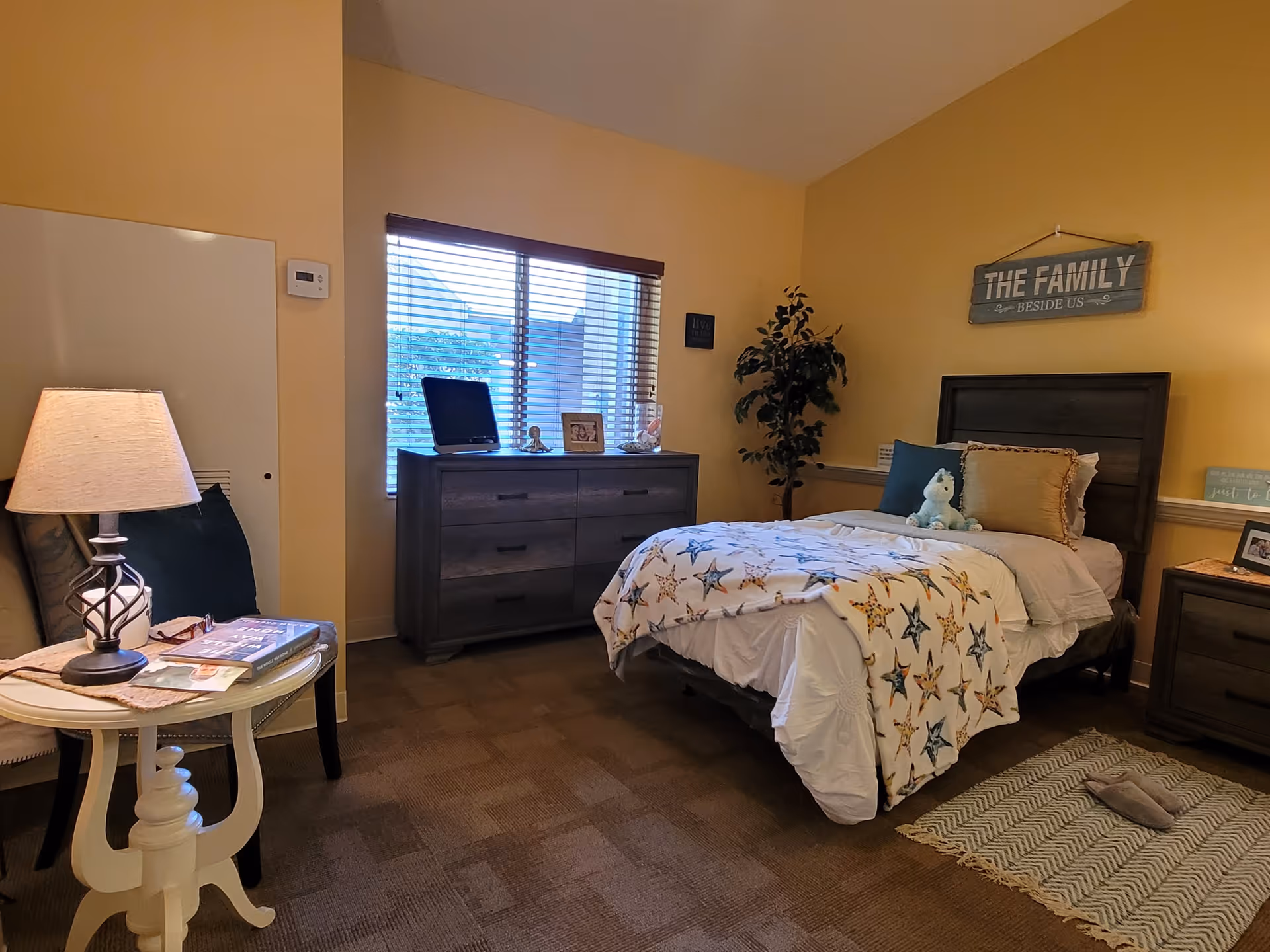 A cozy bedroom with a single bed covered in white bedding and a star-patterned blanket. There are pillows and a stuffed animal on the bed. A wooden dresser with decorative items sits under a window with blinds. A small round table with a lamp and books is next to a chair. The walls are painted yellow, and there is a sign above the bed that reads 'THE FAMILY BESIDE US'. A small plant is in the corner, and a rug is placed on the carpeted floor.