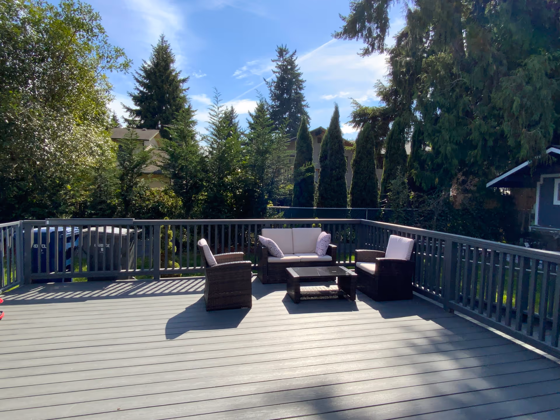 Sunny wooden deck with a sofa, chairs and coffee table surrounded by a railing and tall trees.