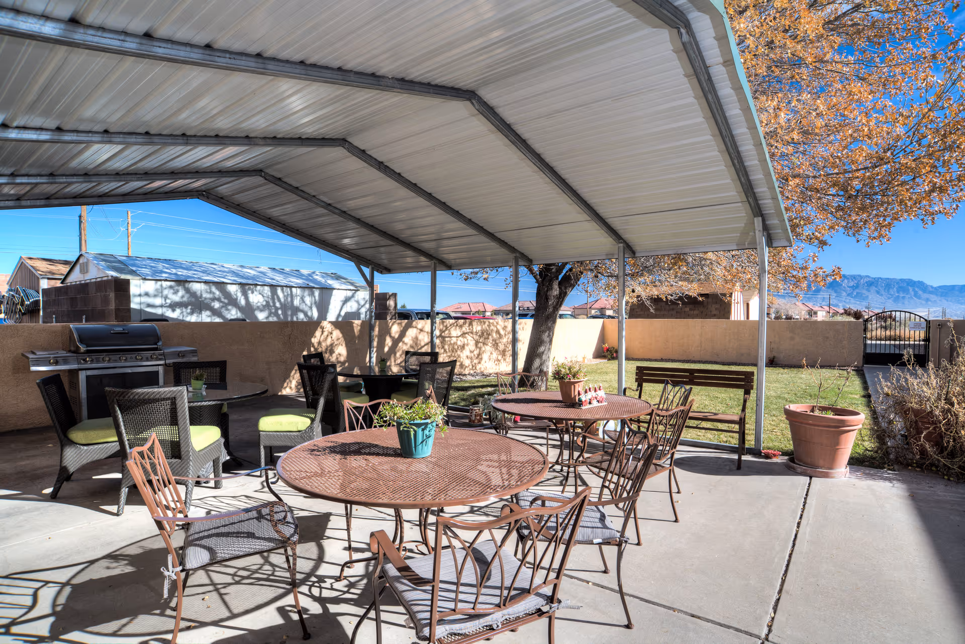 Outdoor covered patio area with several metal tables and chairs, a barbecue grill, potted plants, and a bench. The patio overlooks a grassy yard with a tree and a gated fence, with mountains visible in the background under a clear blue sky.