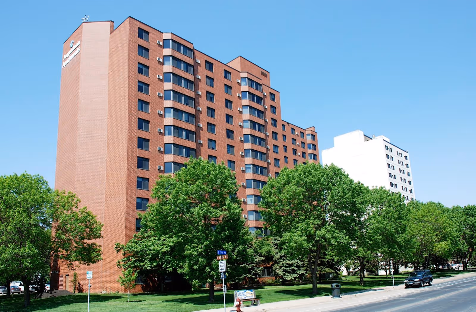 Exterior view of a multi-story brick building with many windows, surrounded by green trees and a clear blue sky. A street runs alongside the building with a few cars parked and driving by. The building has a sign near the top corner.