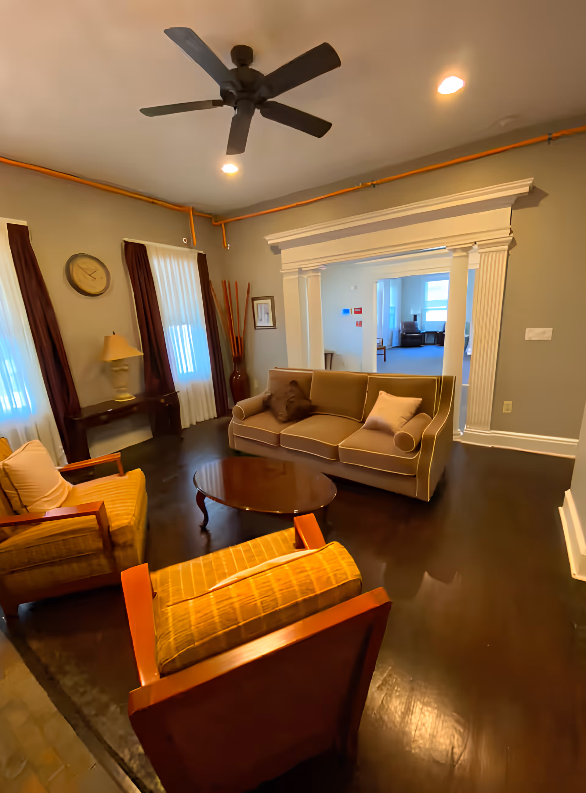 A cozy living room with a beige sofa, two cushioned armchairs, a round wooden coffee table, and a ceiling fan. The room has gray walls, dark wood flooring, two windows with white curtains and dark drapes, a wall clock, a table lamp on a side table, and decorative tall sticks in a vase. There is a wide doorway with white trim leading to another room with more seating and windows.