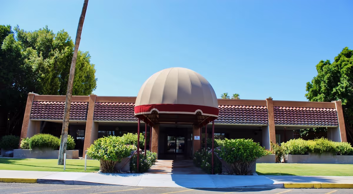 Front exterior view of Rio Vista Post Acute And Rehabilitation facility with a large beige and maroon dome-shaped awning over the entrance, surrounded by green bushes and trees under a clear blue sky.