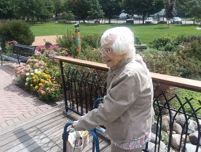 An elderly woman with white hair and glasses using a walker on a wooden deck next to a garden with colorful flowers and greenery. There is a black metal railing along the deck and a park bench on a brick pathway nearby. Trees and a grassy area are visible in the background.