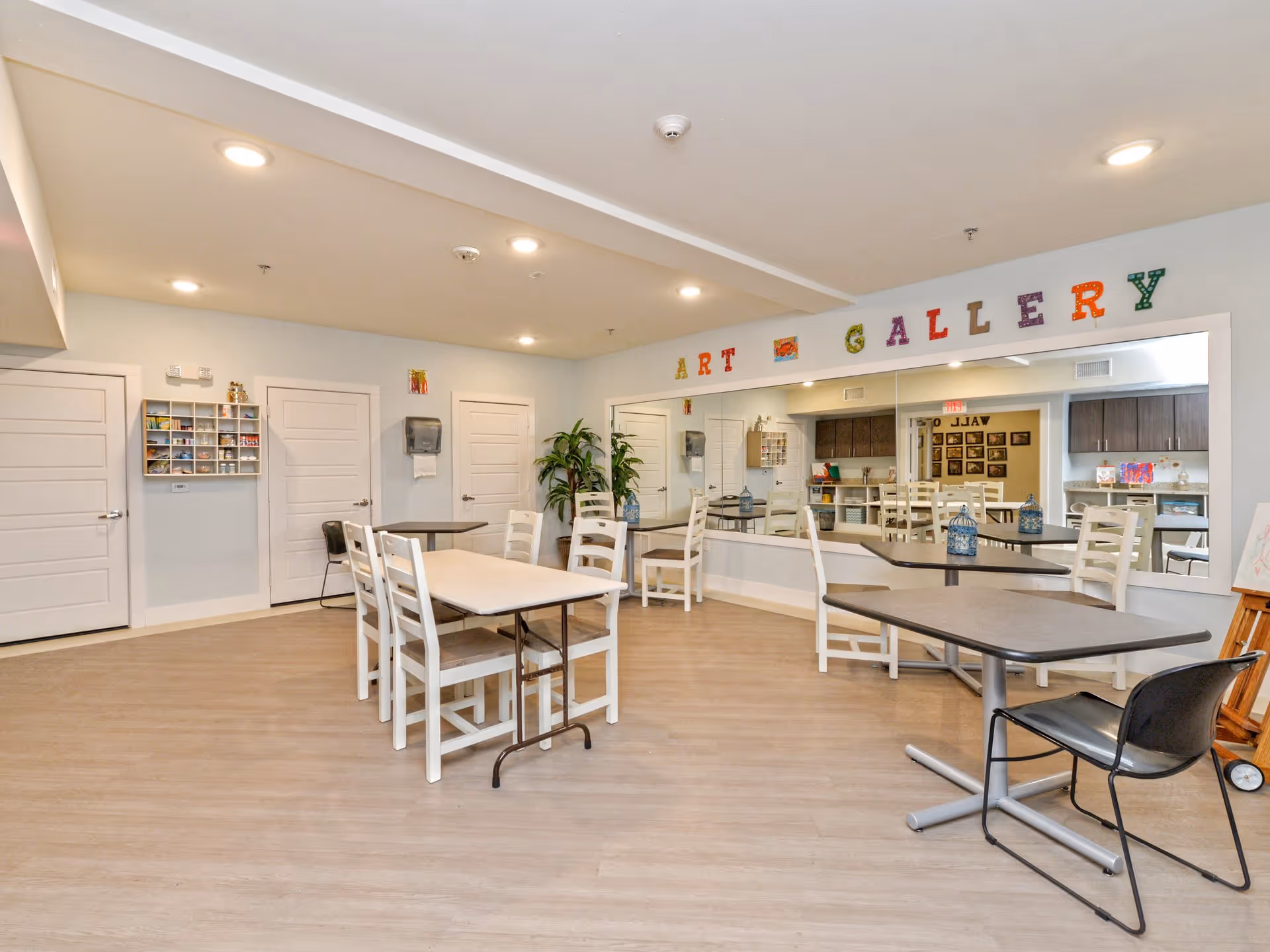 A bright and spacious room with several tables and chairs arranged for group activities. The wall features a large mirror and colorful letters spelling out 'ART GALLERY'. There are doors, a plant, and shelves with art supplies visible in the background.