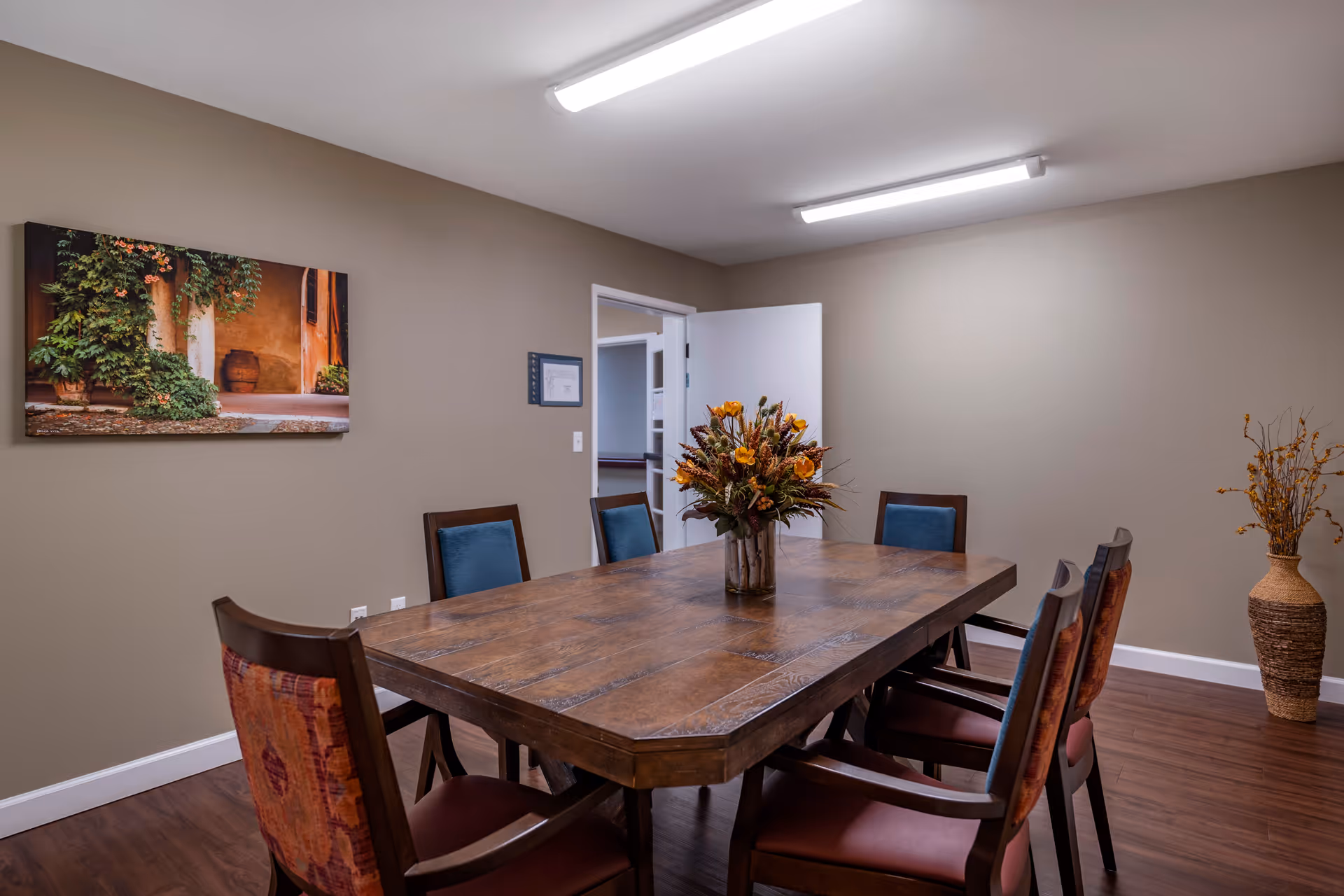 A dining room with a large wooden table surrounded by six chairs, each with a cushioned seat and backrest. A floral arrangement in a vase is placed in the center of the table. The walls are painted beige, with a framed picture of a rustic outdoor scene hanging on one wall. There is a doorway with an open white door leading to another room, and a tall decorative vase with dried branches stands in the corner. The floor is wooden, and the ceiling has fluorescent lighting.