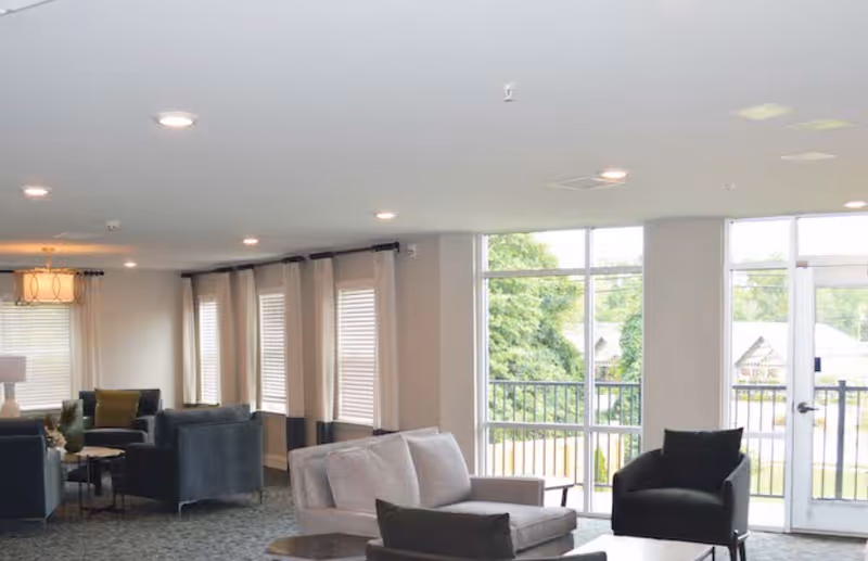 A bright and spacious senior living common area with multiple seating arrangements including gray and black armchairs and sofas. Large windows and glass doors allow natural light to fill the room, offering a view of greenery outside. The room features neutral-colored walls, carpeted floors, and ceiling lights.
