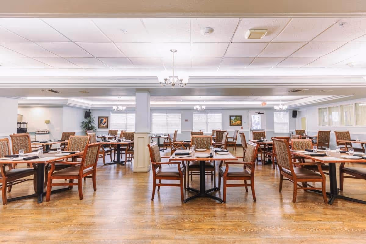 A spacious dining room with multiple wooden tables and cushioned chairs arranged neatly. Each table is set with placemats, napkins, and glasses. The room has large windows with blinds, light-colored walls, and a wooden floor. Ceiling lights and chandeliers provide illumination.