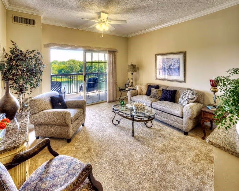 A bright living room with beige walls and carpet, featuring a beige sofa with multiple cushions, an armchair, a glass coffee table with decorative items, a side table with a lamp, and a large sliding glass door leading to a balcony with outdoor chairs and a view of trees and water.