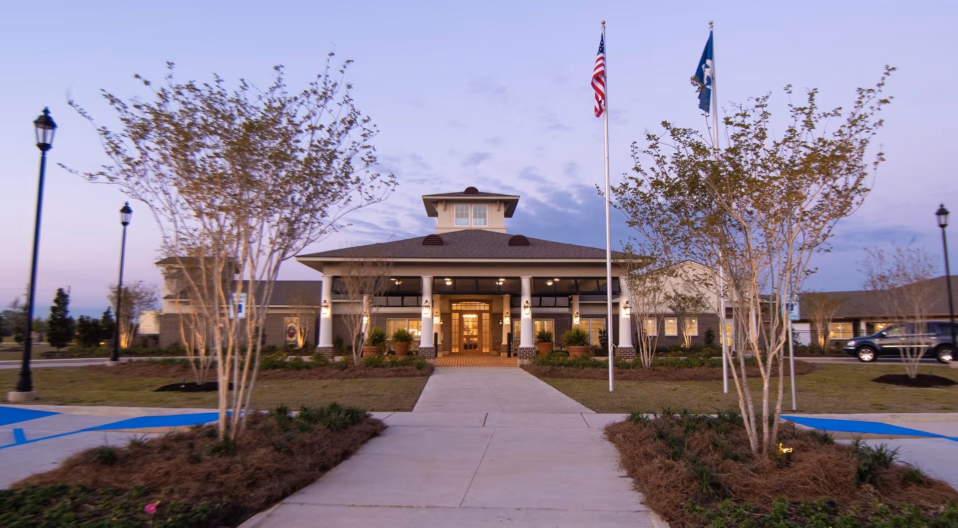 Front exterior view of Cypress Point Nursing & Rehabilitation Center, LLC at dusk, showing a wide walkway leading to the entrance with two flagpoles displaying the American flag and another flag, surrounded by landscaped trees and lamp posts.