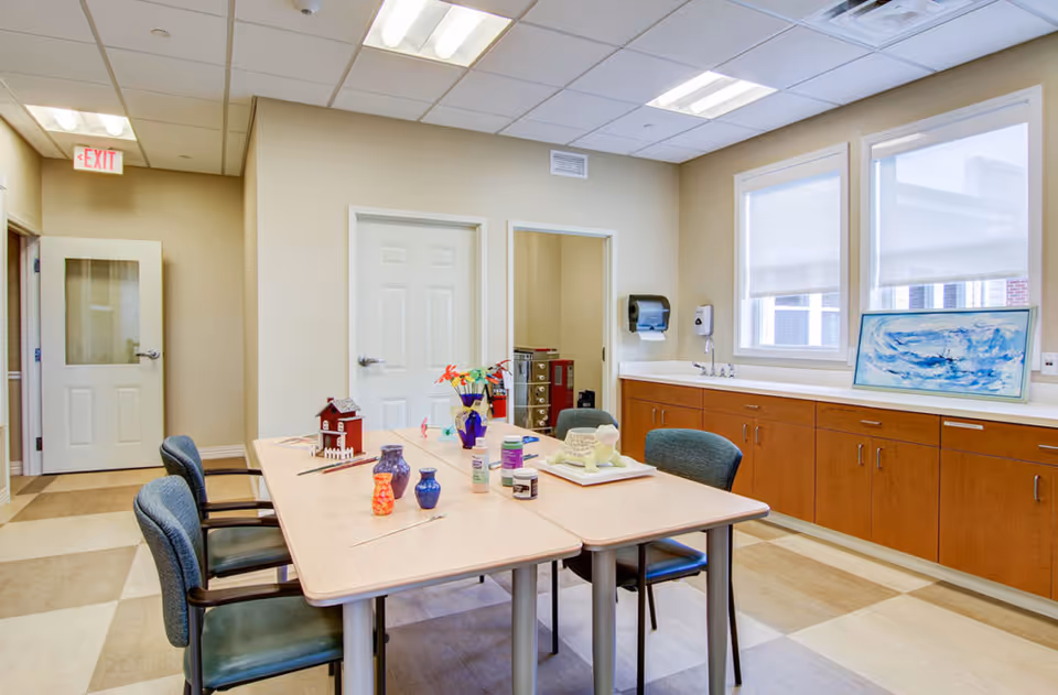 A bright room with a rectangular table surrounded by four chairs. On the table are various craft supplies including paint bottles, paintbrushes, a small model house, and a sculpture. The room has large windows with white blinds, wooden cabinets with a countertop, a sink, and a paper towel dispenser. There are two closed doors and an exit sign above one door.