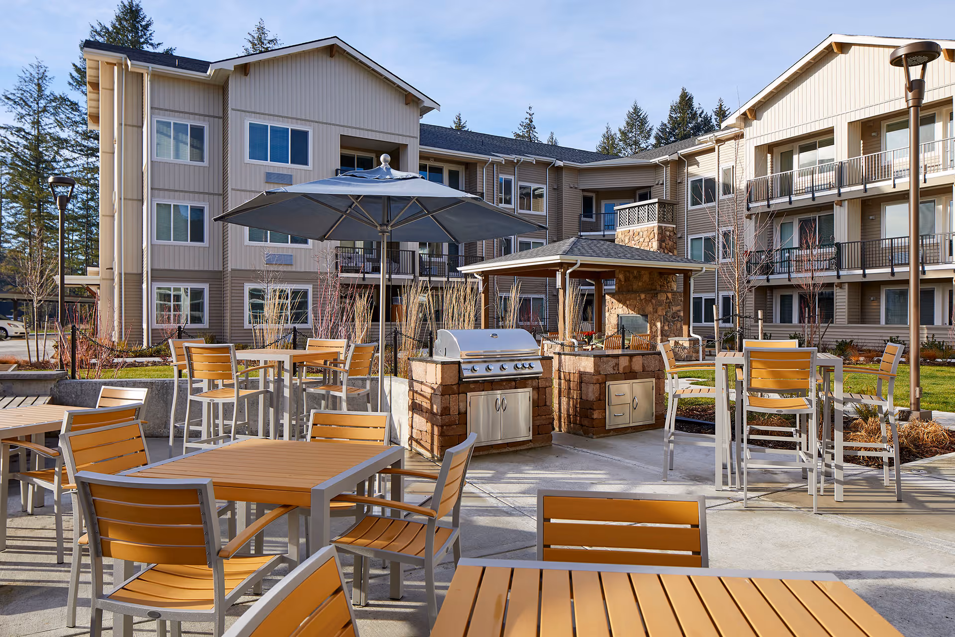 Outdoor patio area at Revel Lacey featuring multiple wooden tables and chairs with metal frames, a large umbrella, and built-in stone grills. The background shows a three-story residential building with balconies and windows surrounded by trees and landscaping.