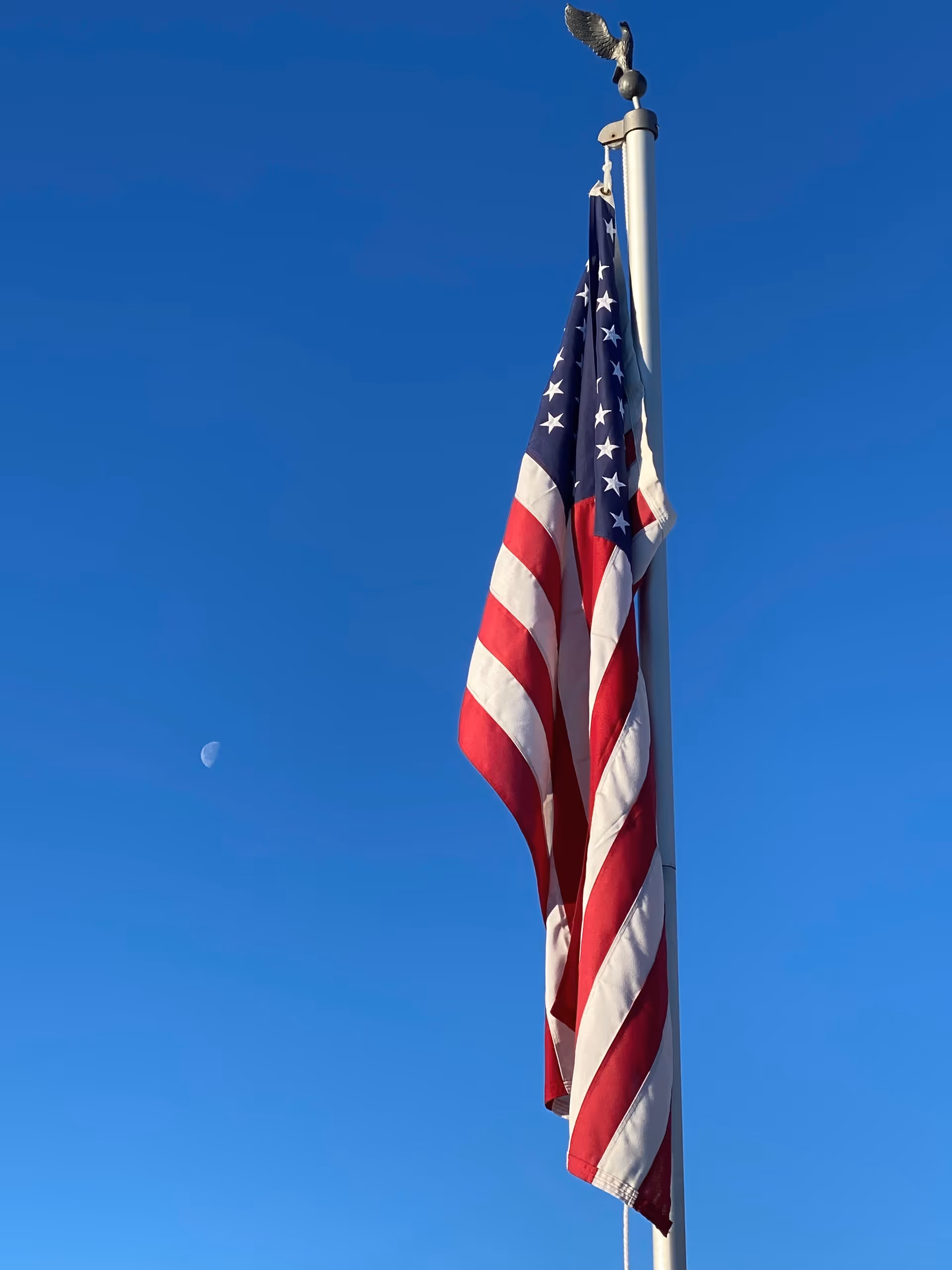 An American flag hanging on a flagpole with a clear blue sky and a visible moon in the background.